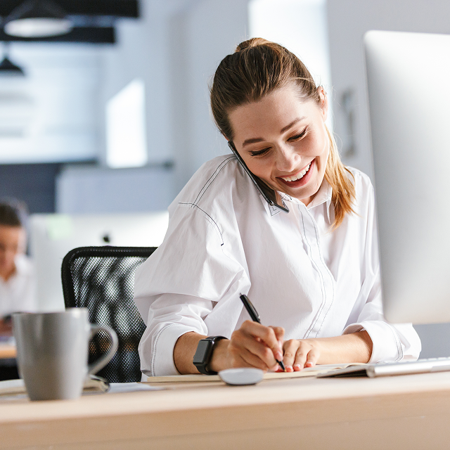 Smiling woman in a crisp white shirt sitting at her desk while speaking to a customer on the phone and writing a note in her notebook