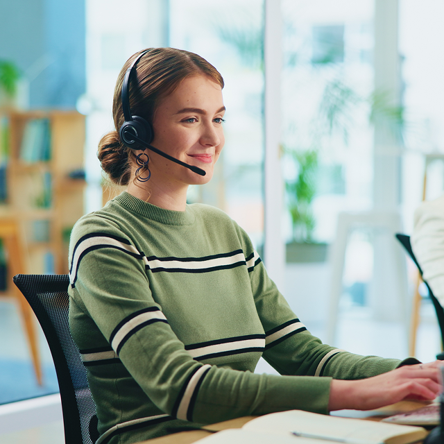 Smiling customer support agent in a striped green jumper sitting at her desk and speaking to a client over her headset